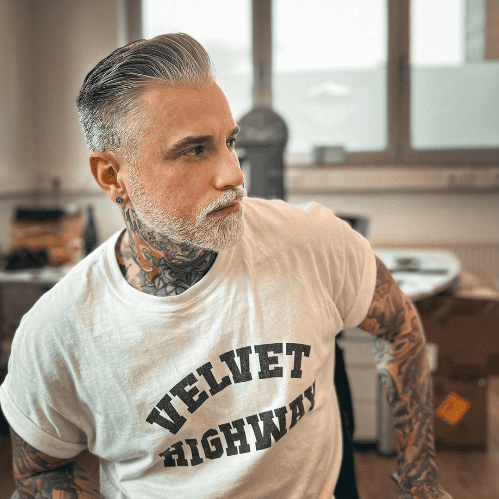 Man with a salt and pepper slicked back haircut, featuring neatly groomed hair with a blend of gray and natural tones. He has a matching beard and visible tattoos, wearing a white t-shirt that reads "Velvet Highway." This image captures a stylish and sophisticated variation of the slicked back hairstyle, perfect for mature men in 2024 seeking a distinguished and modern look.