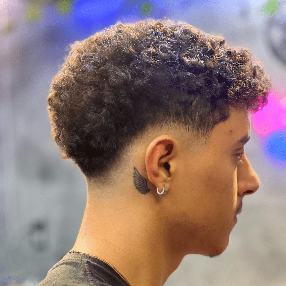 Man with a short fade haircut featuring short curls and a mid drop fade. The hairstyle showcases a clean mid drop fade on the sides and back, blending seamlessly into the natural, textured curls on top. This stylish and modern look is perfect for men's hairstyles in 2024, highlighting the volume and texture of curly hair.