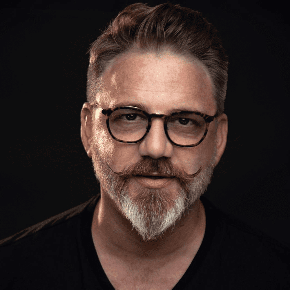 Portrait of a man with a short ducktail beard and curled mustache, wearing round black-rimmed glasses and a black shirt, set against a dark background that accentuates the sharp, stylish beard and mustache grooming.