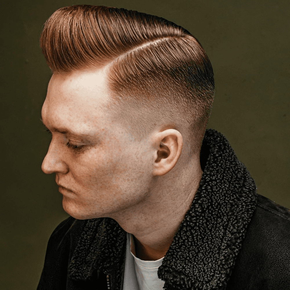 A man with a classic side part pompadour haircut is featured in this image. His hair is neatly parted to one side, with the top section styled into a high, sleek pompadour. The sides are sharply faded, providing a clean and precise look that complements the voluminous top. The crisp parting line adds a touch of elegance, making this hairstyle both timeless and stylish. His expression is serious, reflecting the refined and polished nature of the haircut. The background is simple, keeping the focus on the impeccable hairstyle.