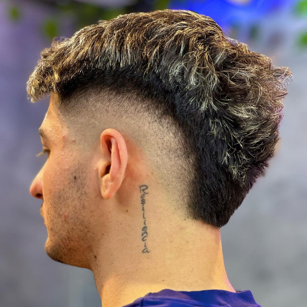 Side profile of a man with a Skin Burst Fade haircut, featuring a high-contrast transition from longer hair on top to shaved skin on the sides and back. The hair on top is styled with texture and volume, while the skin fade creates a sharp and clean look. The haircut also highlights a tattoo with the word "Resiliencia" on the side of the neck. The setting has a blurred background, emphasizing the sharpness and precision of the fade. This skin burst fade is perfect for those seeking a bold, modern, and edgy hairstyle.