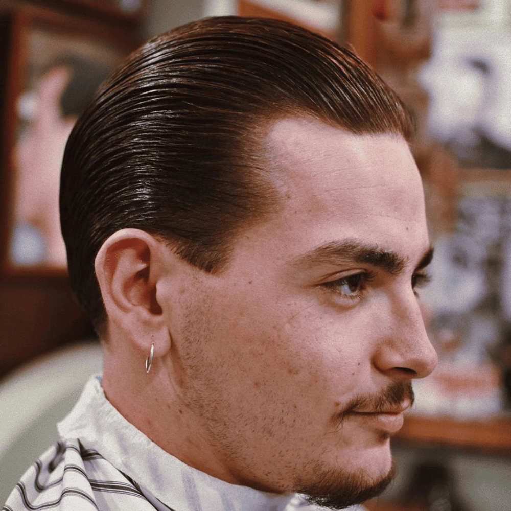 Side profile of a man with a sleek and classy slicked back haircut, featuring smooth, polished hair combed straight back. He is wearing a barber cape, indicating a fresh haircut, and has a small hoop earring and a well-groomed mustache. This image showcases a timeless and refined variation of the slicked back hairstyle, perfect for men in 2024 seeking a sophisticated and elegant look.