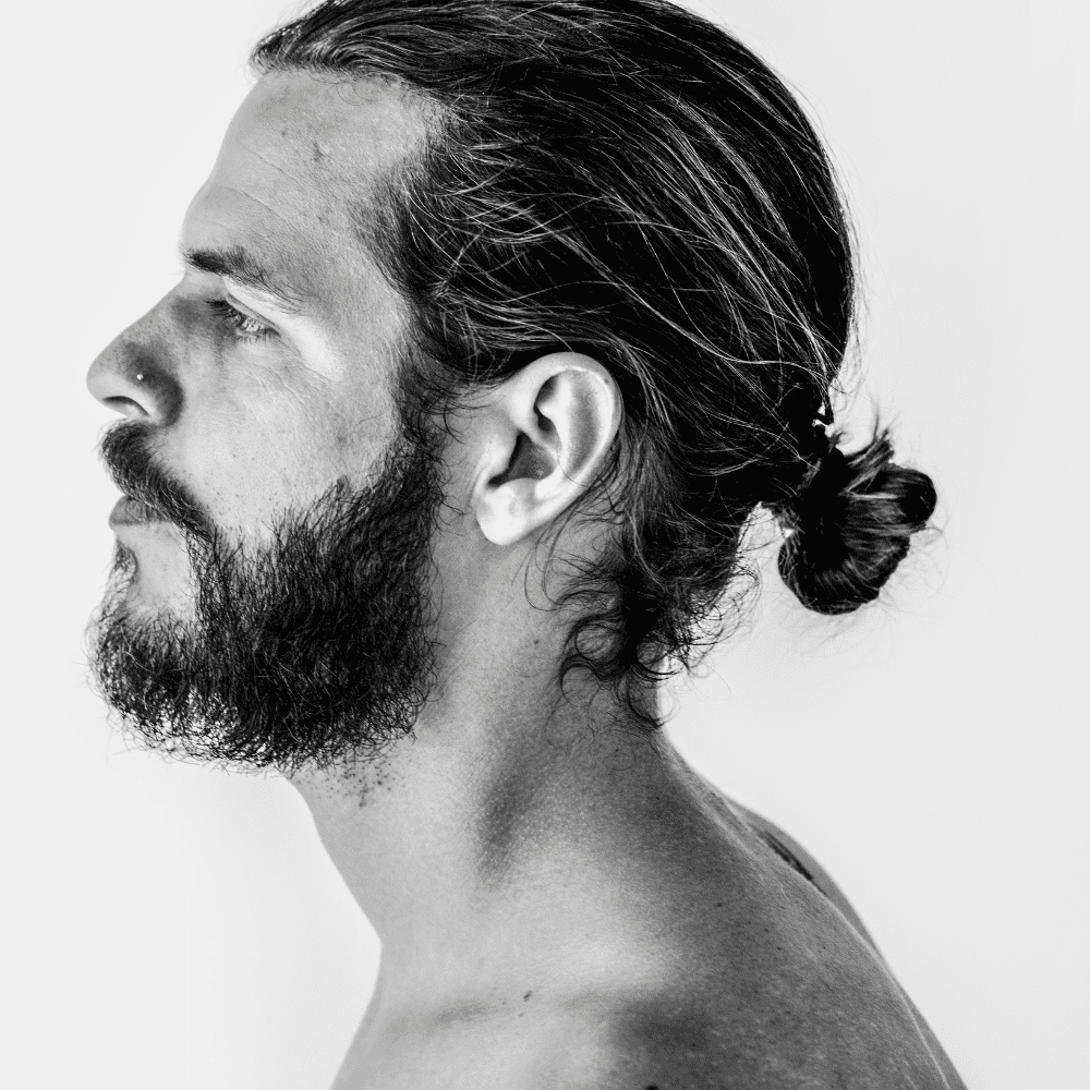 Black and white side profile of a man with a slicked back man bun, featuring long hair pulled back into a bun and a full beard. He has a nose piercing and is shirtless, emphasizing the relaxed yet stylish look. This image captures a modern and casual variation of the slicked back hairstyle, perfect for men in 2024 seeking a laid-back yet fashionable appearance.