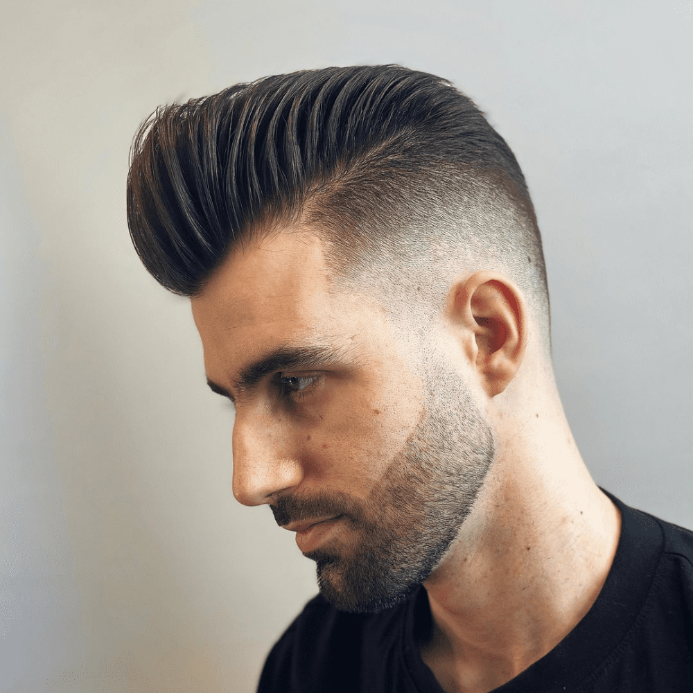 Side profile of a man with a slicked back pompadour haircut, featuring voluminous, high-styled hair on top and a gradual fade on the sides. He has a neatly trimmed beard, enhancing the modern and stylish look. This image showcases a sophisticated and trendy variation of the slicked back hairstyle, perfect for men in 2024 seeking a bold and polished appearance.