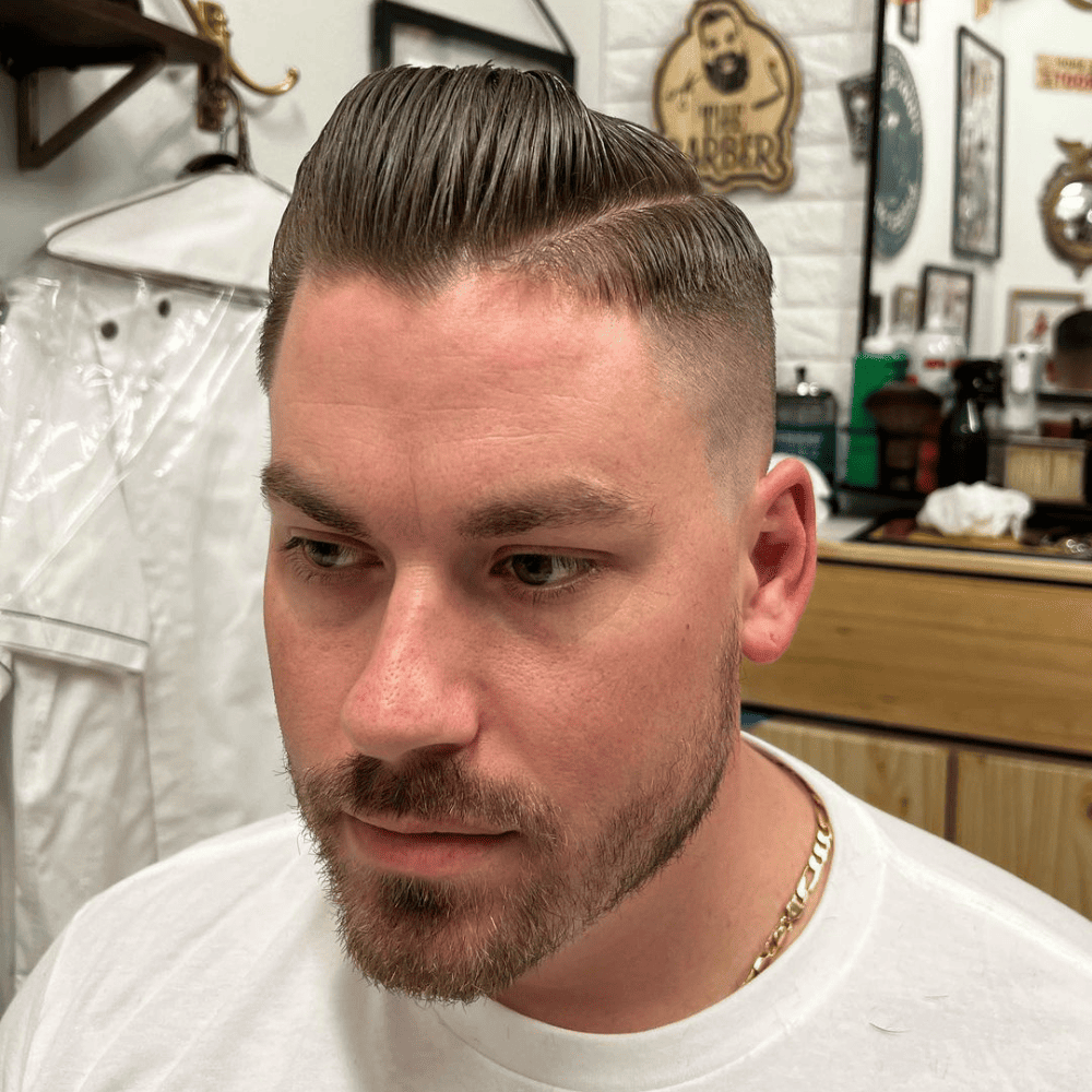 Man with a slicked back haircut featuring a side parting, creating a structured and polished look. He has a neat beard and is seated in a barbershop setting, highlighting the clean lines and classic style of this haircut. This image showcases a sophisticated variation of the slicked back hairstyle, perfect for men in 2024 seeking a timeless and refined appearance.