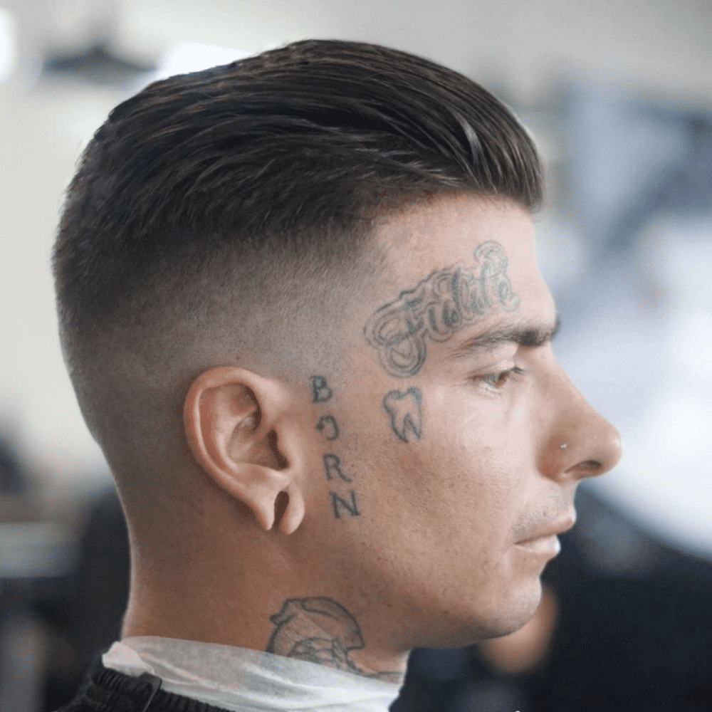 Side profile of a man with a slicked back high fade haircut, featuring tattoos on his face and neck. The hairstyle showcases neatly slicked back hair on top with a clean, high fade on the sides and back, creating a sharp and modern look.