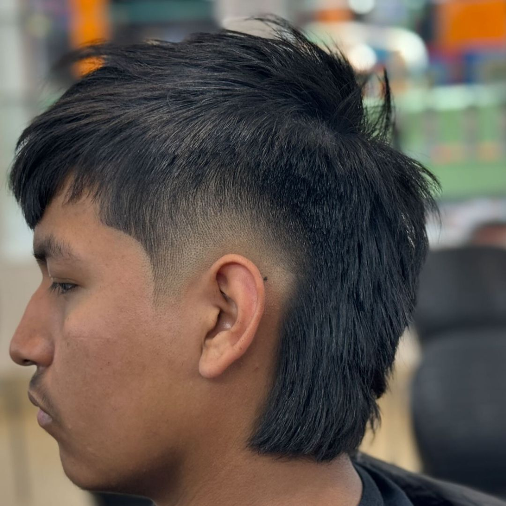 Side profile of a young man with a Straight Hair Mohawk haircut. The hairstyle features straight hair styled into a Mohawk with a high fade on the sides, creating a clean and sharp contrast. The Mohawk extends from the front to the back, with the back lengthened into a subtle mullet style. The man has a smooth, straight hair texture, and the haircut highlights the precision of the fade and the sleekness of the straight hair. The background shows an indoor setting, likely a barbershop, focusing on the modern and edgy look of the Straight Hair Mohawk.