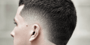 Side profile of a man with a stylish burst fade haircut. The hairstyle features a sharp, clean fade that gradually tapers down the sides and back, creating a rounded 