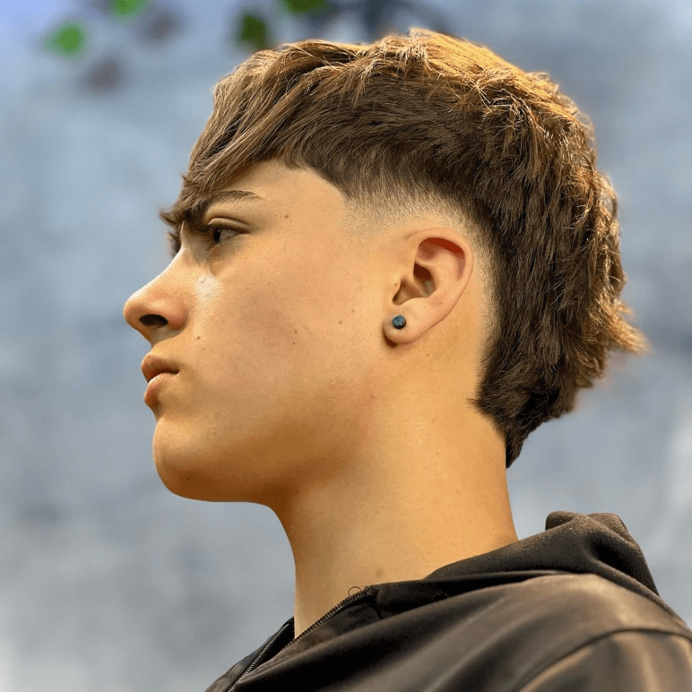 Side profile of a young man with a Taper Mohawk haircut. The hairstyle features a gradual taper fade on the sides, blending seamlessly into the longer hair on top. The top hair is styled with texture and volume, creating a modern, edgy look. The man is wearing a black hoodie and has an earring, with a background that suggests an outdoor setting. The Taper Mohawk combines a clean, tapered fade with a stylish Mohawk, highlighting a balanced and trendy haircut.