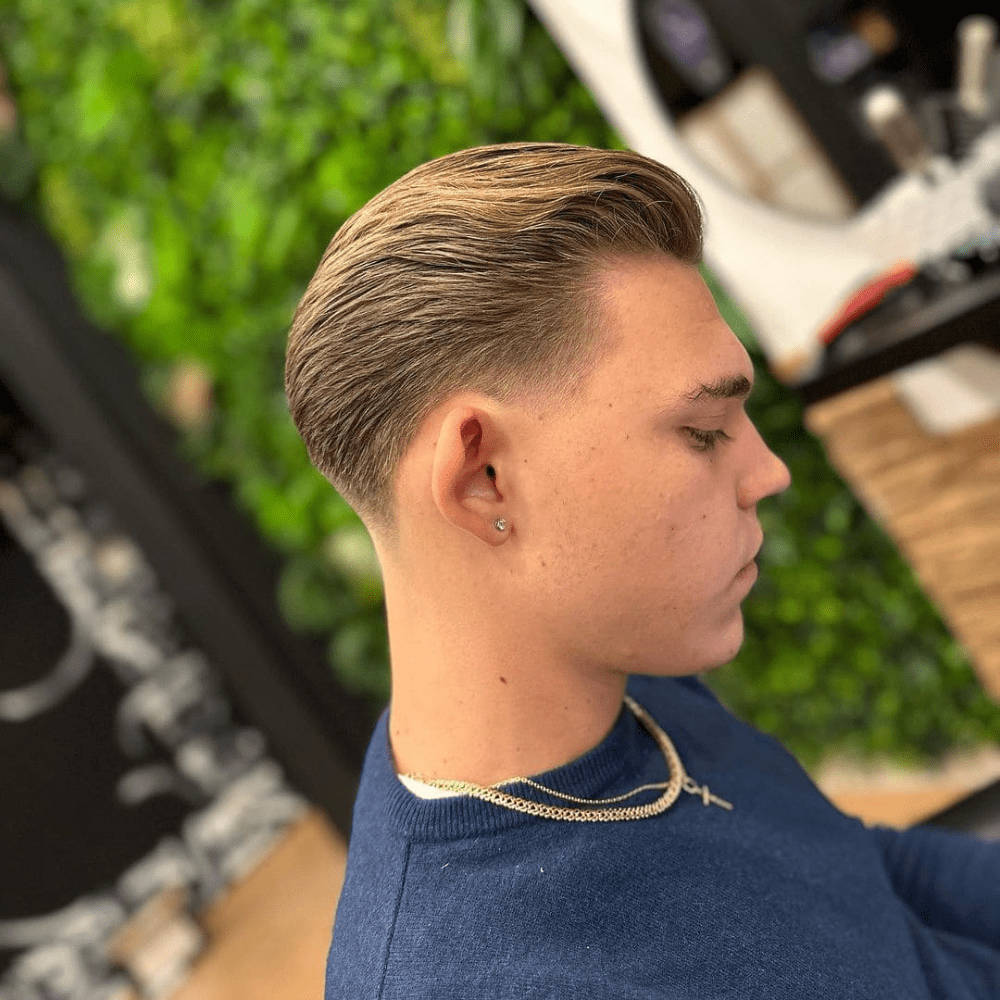 Side profile of a young man with a textured slicked back haircut, featuring a smooth transition from the longer, slicked back top to the shorter sides. He is wearing a blue sweater, a gold chain necklace, and has a small earring, enhancing the modern and stylish look. This image highlights a clean and contemporary variation of the slicked back hairstyle, perfect for men in 2024 seeking a polished and fashionable appearance.