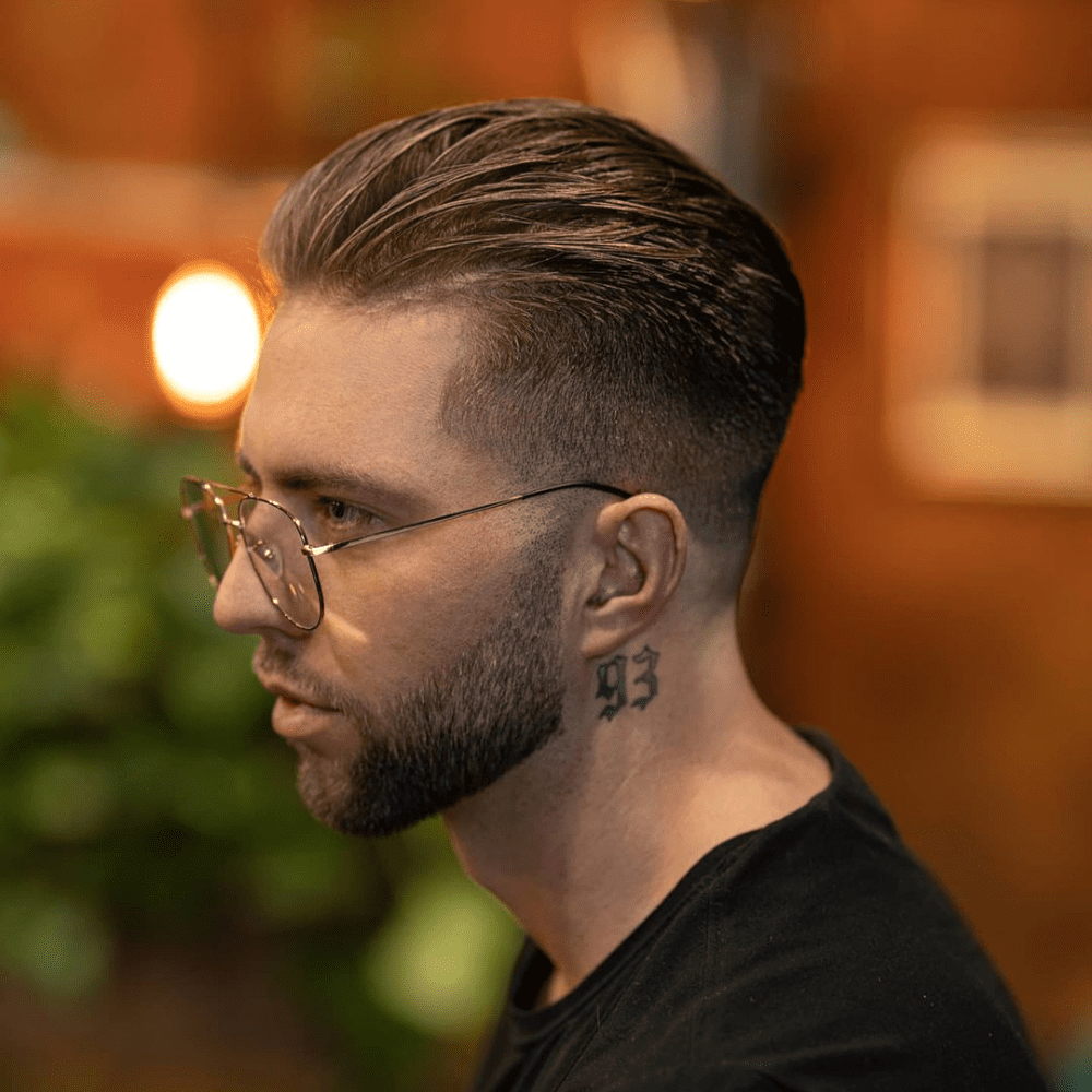 Side profile of a man with a tapered slicked back haircut, featuring a gradual fade from the longer, slicked back top to the shorter sides. He is wearing glasses and has a neck tattoo with the number "93," paired with a neatly trimmed beard. This image showcases a clean and modern variation of the slicked back hairstyle, perfect for men in 2024 seeking a refined and contemporary look.