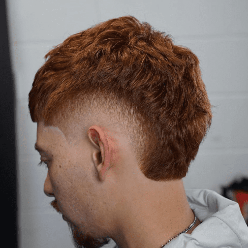 Side profile of a man with a Textured Burst Fade haircut, featuring rich, auburn hair styled with texture and volume on top. The burst fade starts high near the temples and gradually tapers down, creating a seamless blend that accentuates the textured layers. The hair is slightly longer on top, allowing for a dynamic and stylish appearance. The setting includes a neutral indoor background, keeping the focus on the intricacy and sharpness of the haircut. This textured burst fade is perfect for men seeking a modern, fashionable look that showcases natural hair texture.