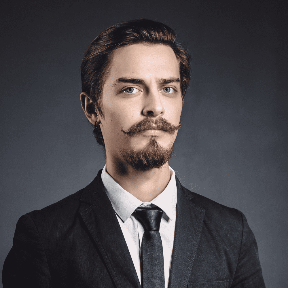 Young man with an Anchor beard and styled mustache, wearing a black suit and tie, posing confidently against a dark grey background. Exemplifying popular beard styles for men, this look highlights precision grooming and sophistication.