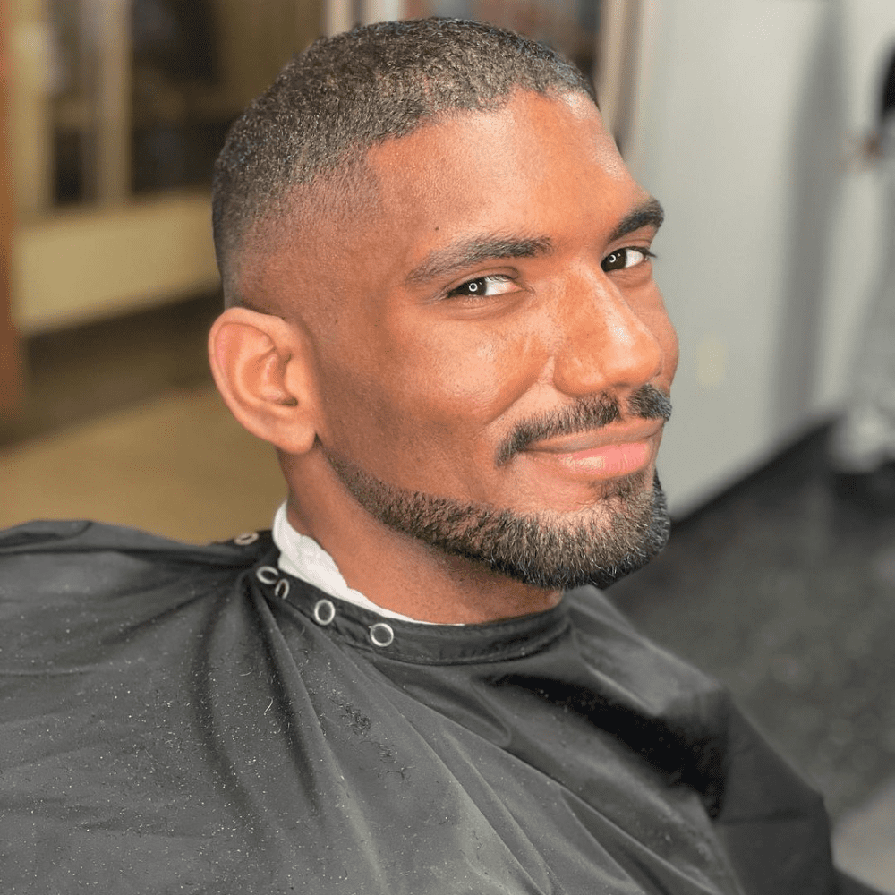 Smiling man with a neatly groomed Balbo beard, sitting in a barber's chair with a cape, showcasing precise facial hair styling. Ideal example of popular beard styles for men, highlighting clean lines and professional grooming.