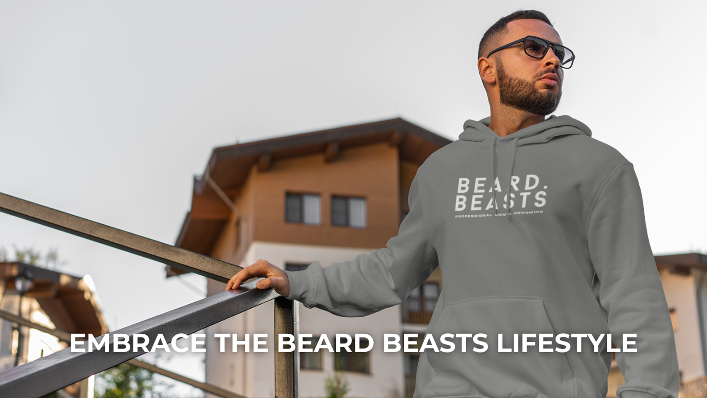 A confident man wearing a gray Beard Beasts hoodie with the logo 'Professional Men's Grooming,' standing near a modern building and exuding style and sophistication.