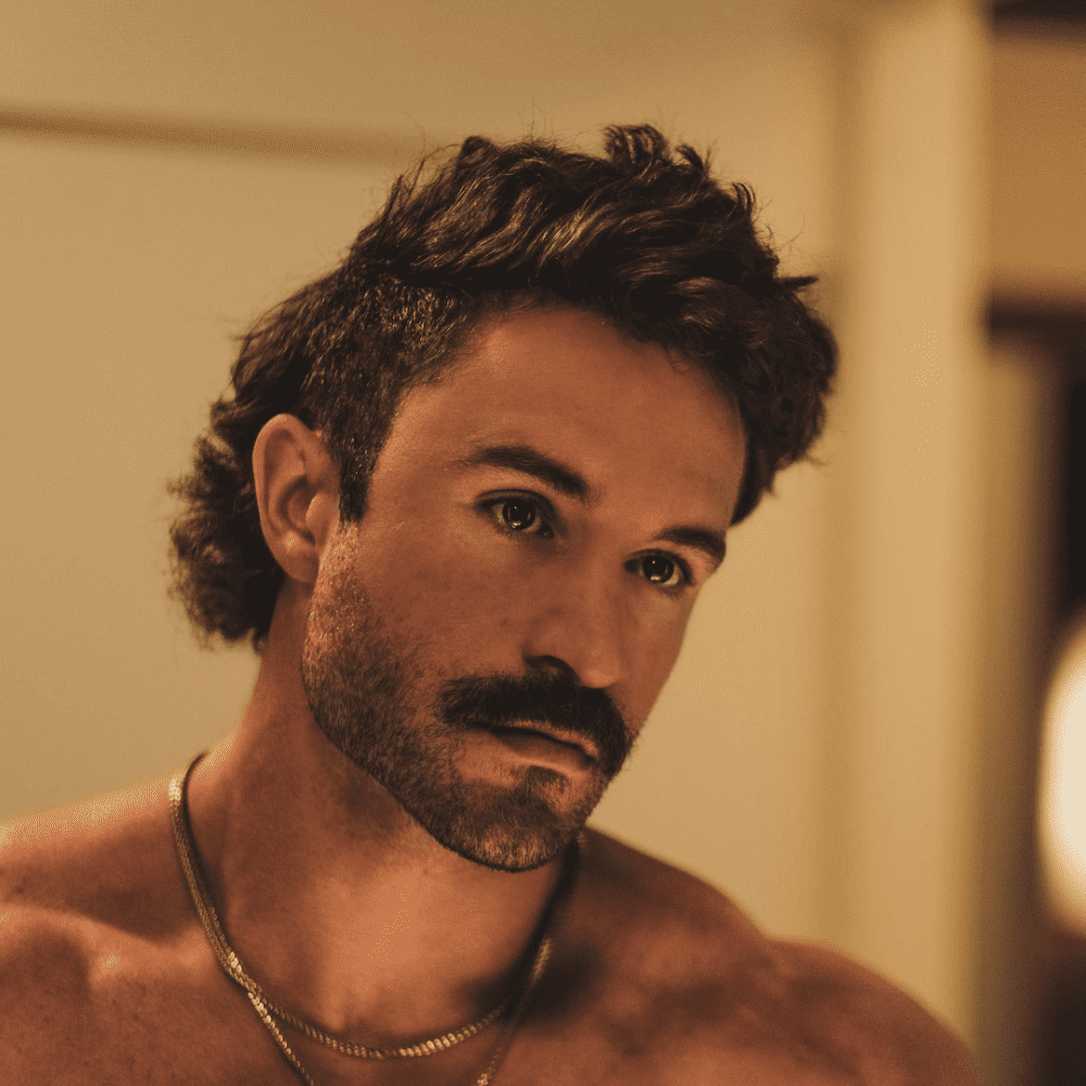 Man with a Beardstache featuring a prominent mustache and short beard, standing shirtless in a warmly lit room, looking contemplative. Highlighting one of the popular beard styles for men, this look blends ruggedness with a touch of refinement.