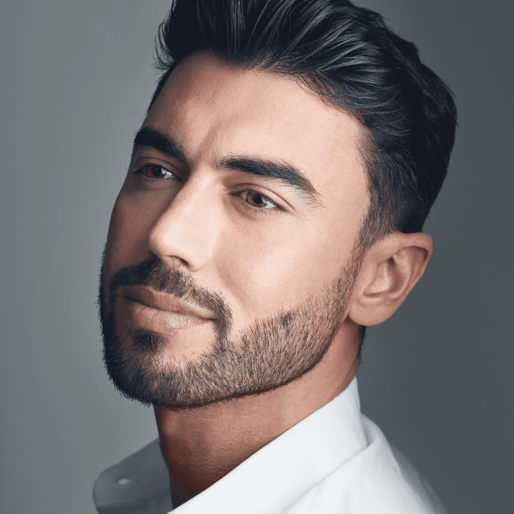 Handsome man with a well-groomed stubble beard, wearing a white shirt, looking off-camera with a confident expression. Showcasing one of the popular beard styles for men, this look combines simplicity with a touch of rugged elegance.