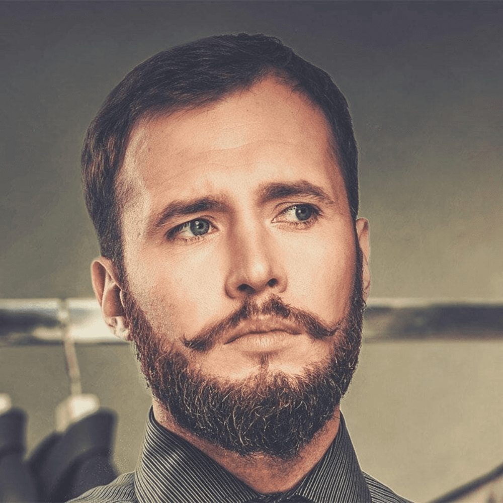 Man with a Verdi beard featuring a styled mustache, wearing a striped dress shirt, looking thoughtfully into the distance. Showcasing one of the popular beard styles for men, this look blends classic elegance with modern grooming.