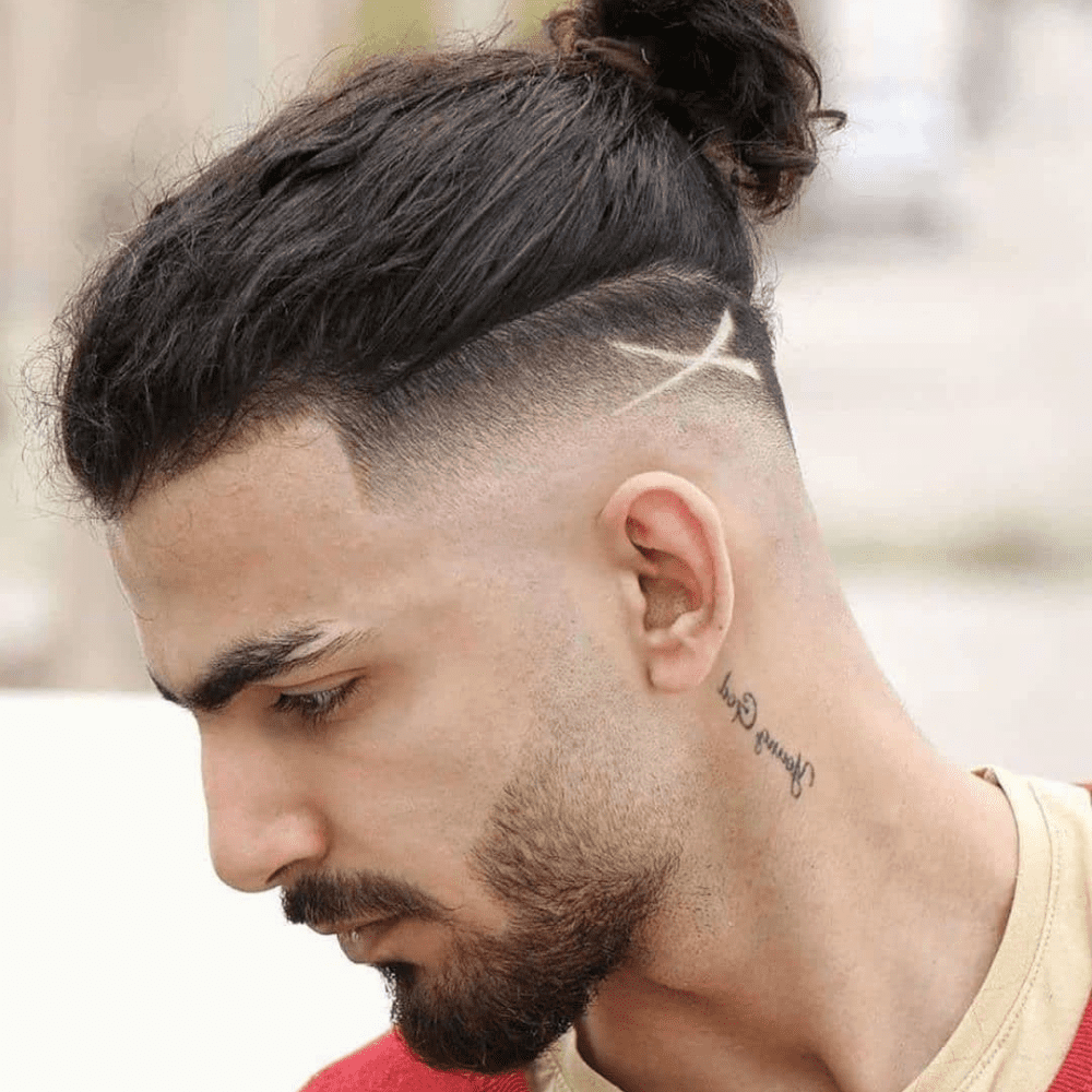 Side profile of a man with an undercut slicked back man bun, featuring shaved sides with a design and long hair gathered into a bun. He has a neatly trimmed beard and a neck tattoo that reads "Young God." This image highlights a bold and stylish variation of the slicked back hairstyle, perfect for men in 2024 seeking a modern and edgy look with added flair.