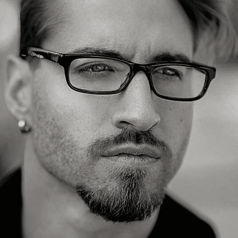 Man with a Van Dyke beard, wearing glasses and an earring, captured in a thoughtful expression. This black and white image highlights one of the popular beard styles for men, showcasing a distinct combination of a mustache and pointed goatee for a refined and artistic look.