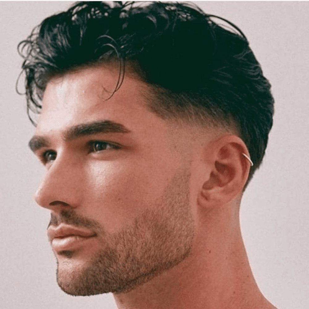 Side profile of a man with a wavy slicked back haircut, featuring natural waves styled back and a clean fade on the sides. He has a neatly groomed beard and an ear piercing, adding to the modern and stylish look. This image showcases a textured and trendy variation of the slicked back hairstyle, perfect for men in 2024 seeking a relaxed yet polished appearance.